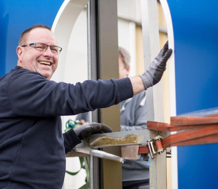 Lächelnder Metallbauer montiert Fenster in einer Baustelle, trägt Arbeitskleidung und Schutzhandschuhe.