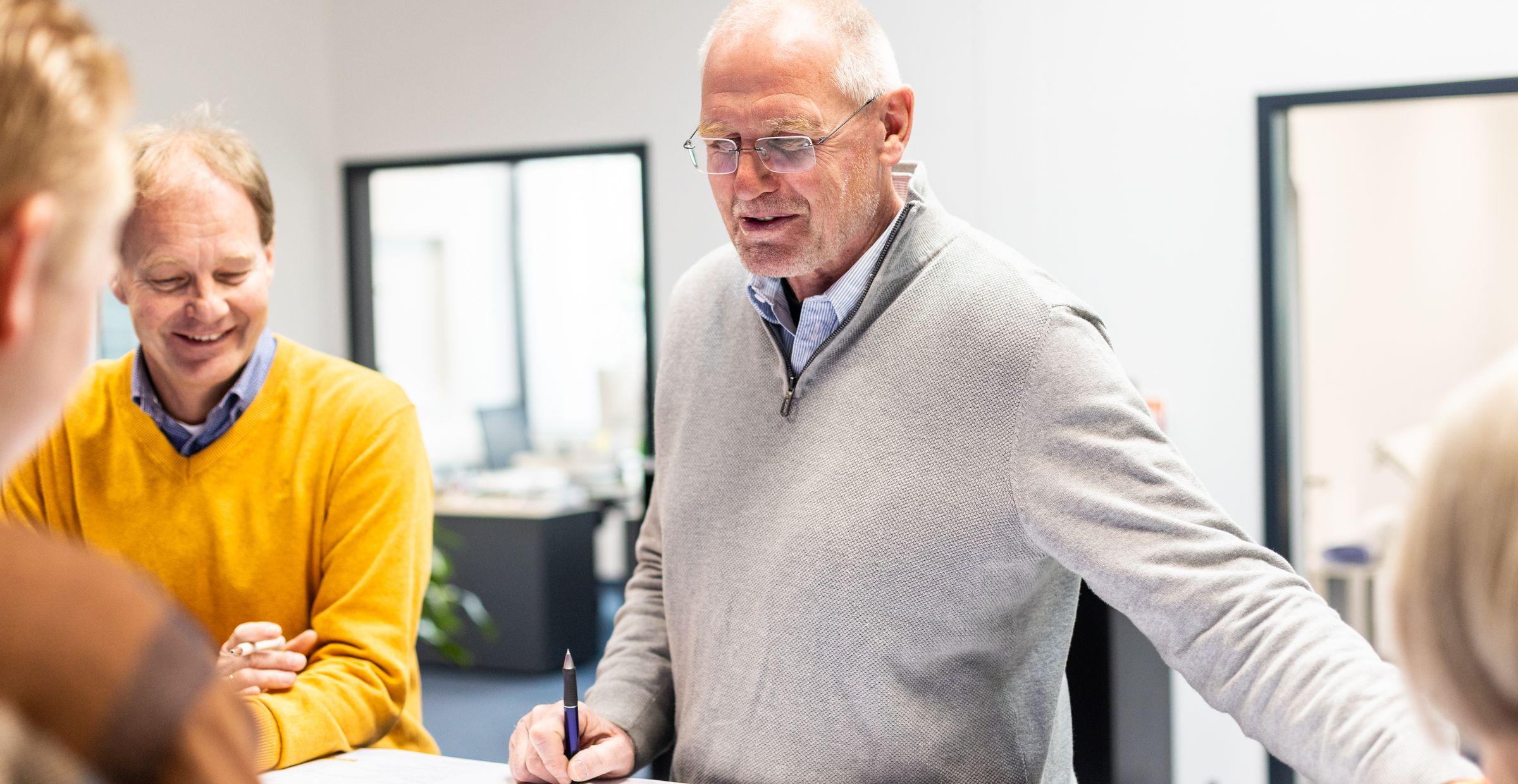 Mehrere Personen stehen um einen Tisch und besprechen technische Zeichnungen. Ein älterer Mann im grauen Pullover erklärt etwas mit einem Stift in der Hand, während ein anderer Mann in einem senfgelben Pullover lächelt. Die Szene wirkt wie eine konstruktive Besprechung im Büro.