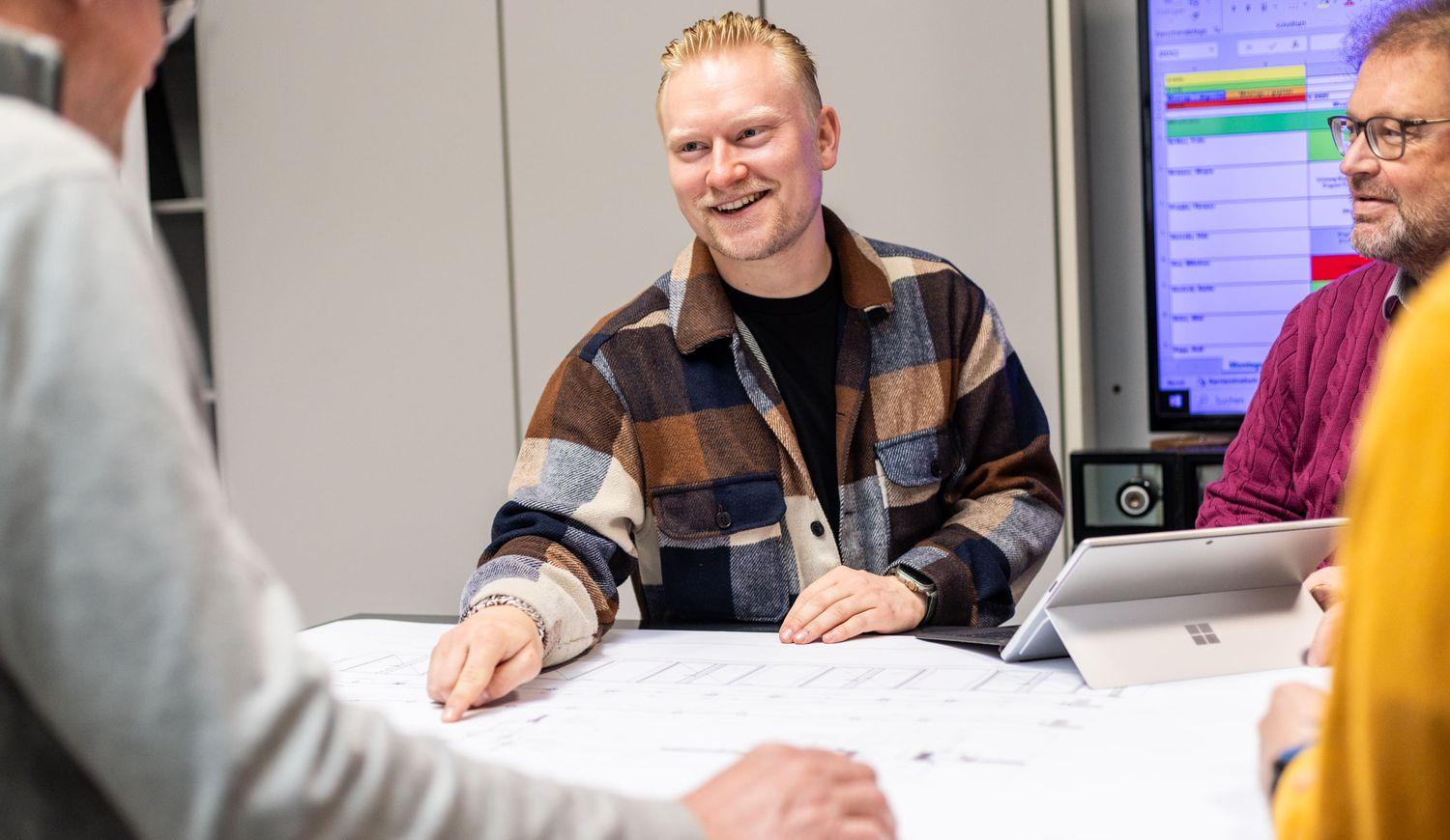 Person mit kariertem Hemd sitzt an einem Tisch mit ausgebreiteten Plänen und spricht mit mehreren Personen in einem Büro mit Bildschirm im Hintergrund