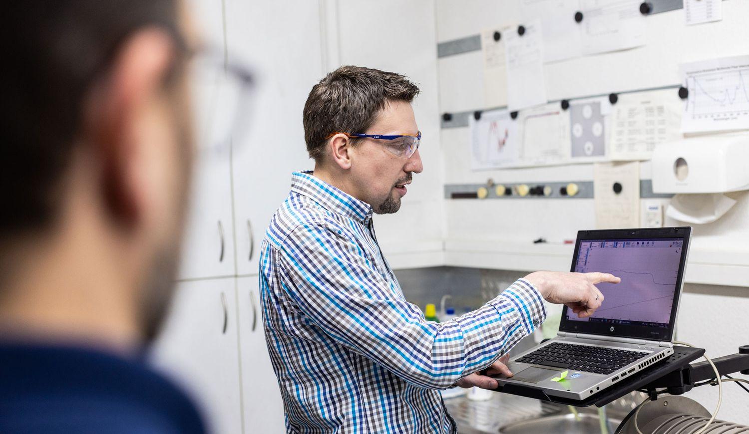 Mann in kariertem Hemd und Schutzbrille arbeitet an einem Laptop in einem modernen Labor mit weißen Schränken und Geräten im Hintergrund