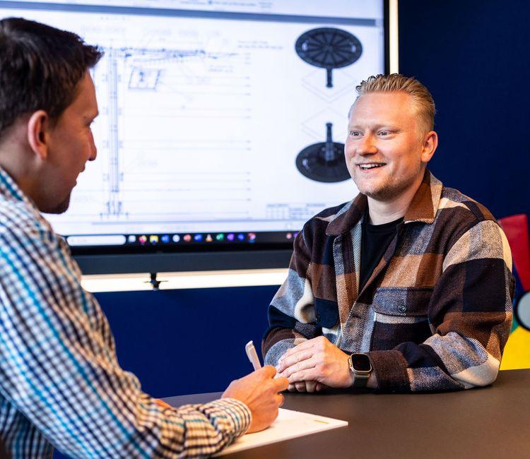 Zwei Männer in einem Gespräch am Schreibtisch - einer in kariertem Hemd, der andere in gemustertem Pullover lächelnd, mit Computer-Bildschirm im Hintergrund